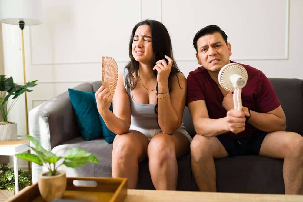 Sweating couple relaxing indoors, escaping scorching summer temperatures by lounging together on comfortable living room sofa