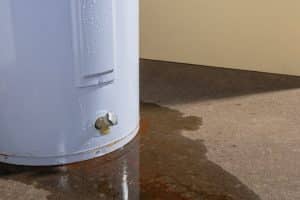 Water leaking from a residential electric water heater sitting on a concrete floor with signs of rust and iron staining the floor.