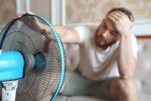 Young bearded man using electric fan at home, sitting on couch cooling off during hot weather, suffering from heat, high temperature