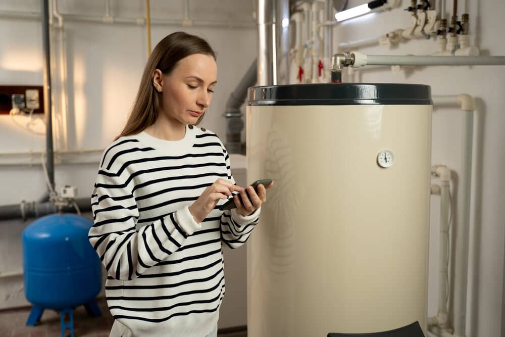 A young woman examines a non-operational boiler with concern, holding a phone in her hand, likely seeking help or planning to call for repair services. The basement setting indicates a residential location where the boiler malfunction requires immediate attention due to cold weather.