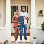 Portrait of a young couple holding a chalkboard with our first home written on it.