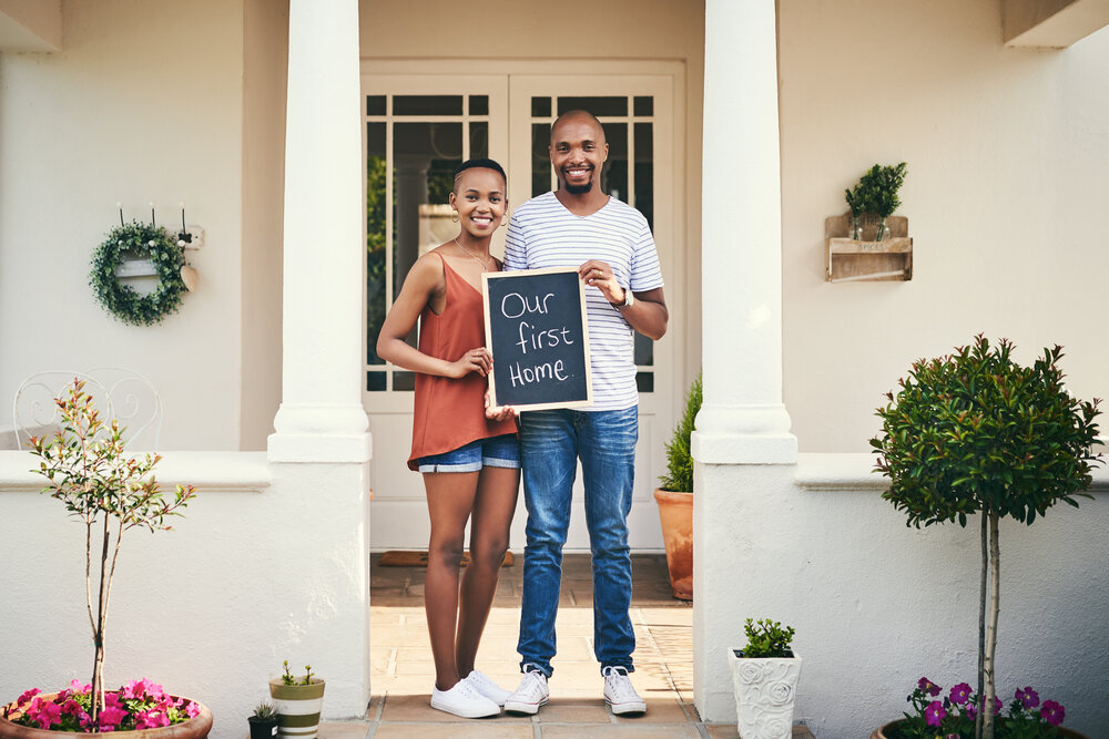 Portrait of a young couple holding a chalkboard with our first home written on it.