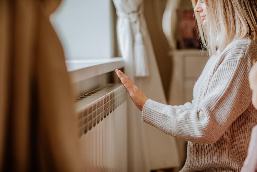 Young blond woman in long winter beige sweater is posing at home near the radiator.