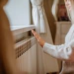 Young blond woman in long winter beige sweater is posing at home near the radiator.