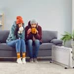 A young man and woman sitting on sofa in the living room in winter outerwear and in hats at home warming their hands from a space heater. Heating problems and broken furnace concept.