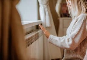 Young blond woman in long winter beige sweater is posing at home near the radiator.