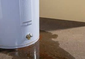 Water leaking from a residential electric water heater sitting on a concrete floor with signs of rust and iron staining the floor.
