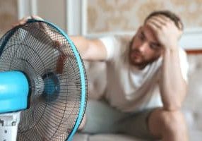 Young bearded man using electric fan at home, sitting on couch cooling off during hot weather, suffering from heat, high temperature