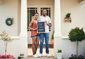 Portrait of a young couple holding a chalkboard with our first home written on it.