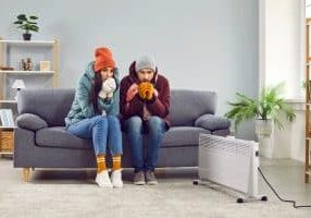 Prepare Your Furnace for Winter A young man and woman sitting on sofa in the living room in winter outerwear and in hats at home warming their hands from a space heater. Heating problems and broken furnace concept.